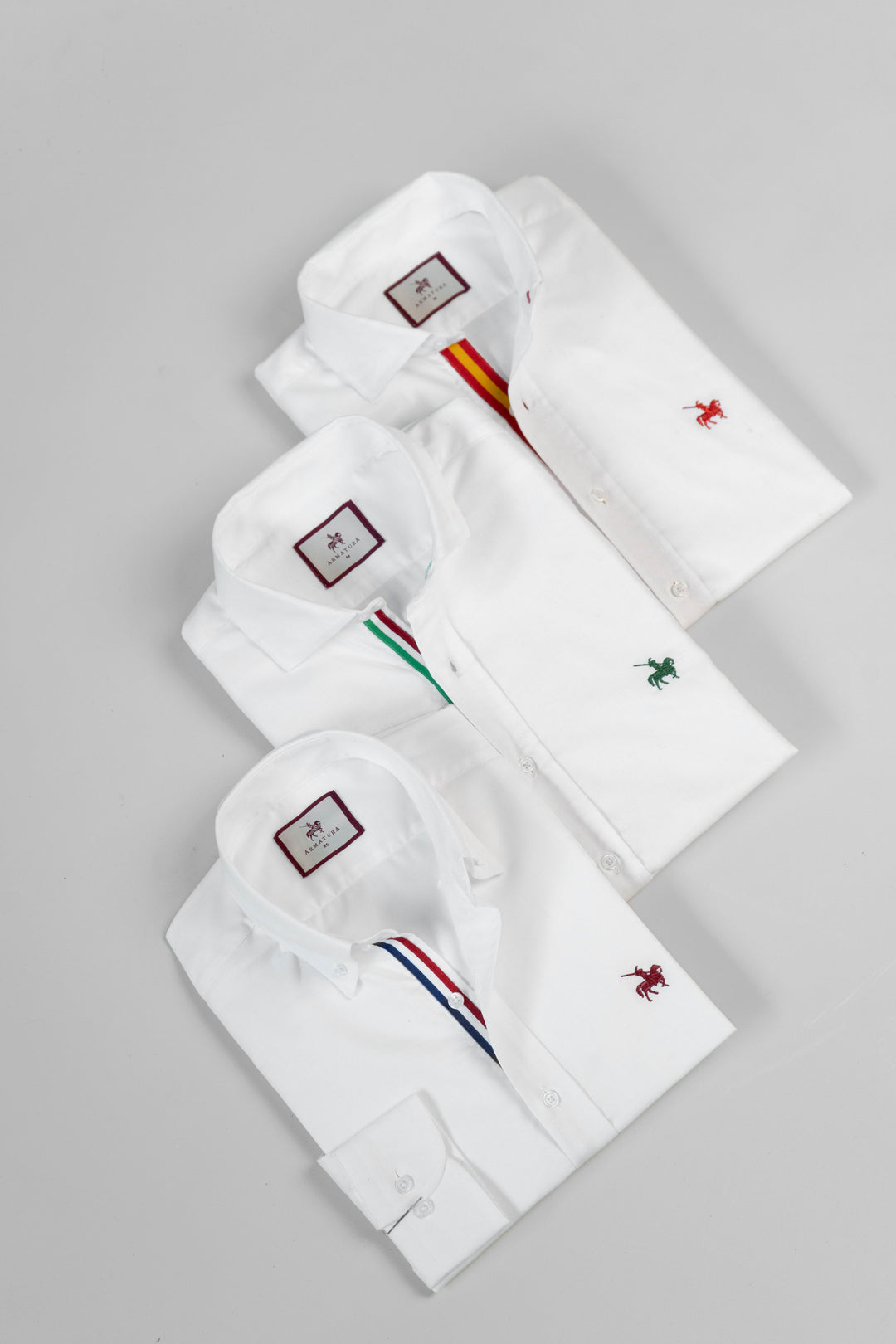 3 camisas blancas para hombre Insignia Armatura. Camisas manga larga con detalles de colores en el cuello y logo bordado en el pecho de distintos colores. Imagen producto expuesto 3 camisas.